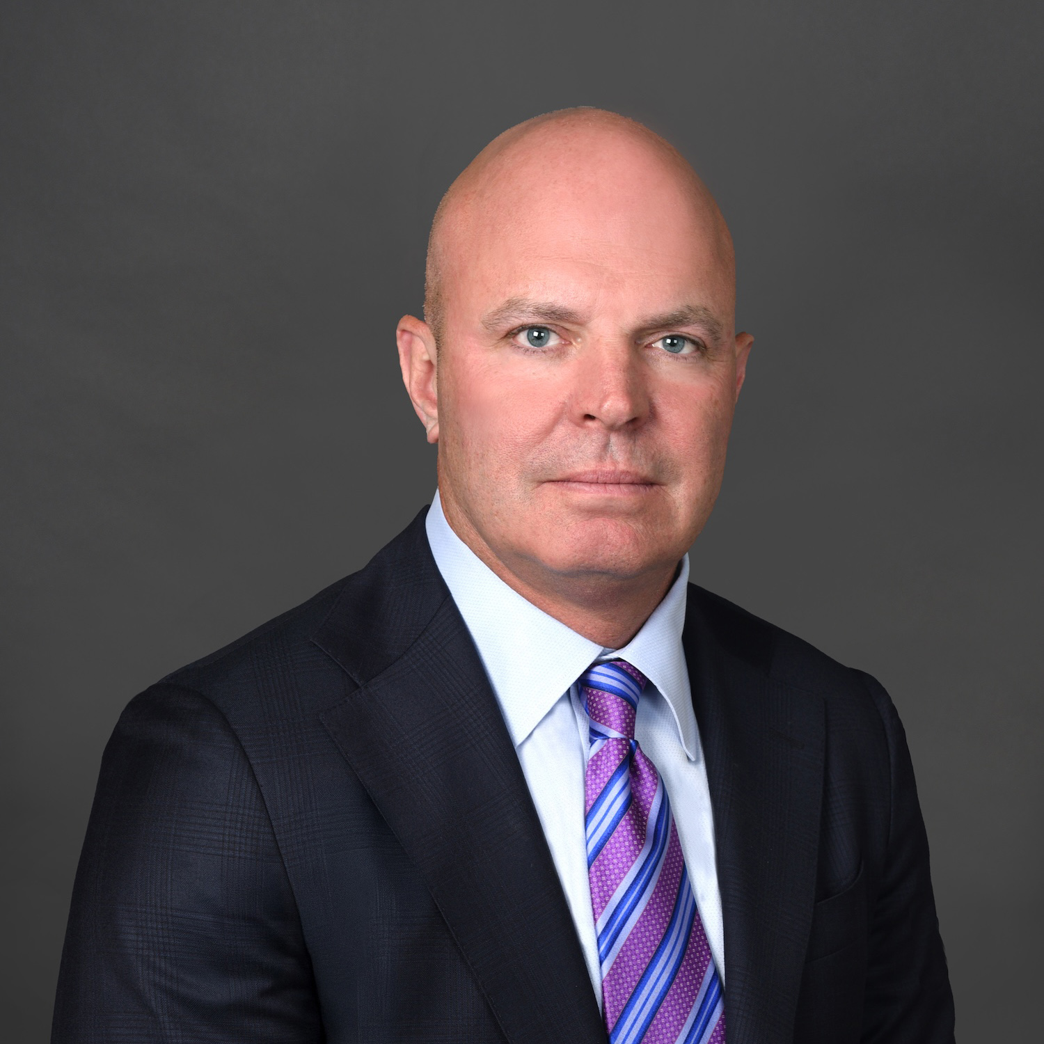 A bald man in a dark suit, light blue shirt, and purple striped tie poses against a plain gray background, looking directly at the camera with a neutral expression.